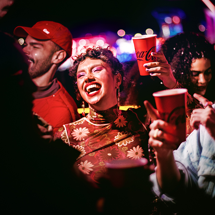 Un grupo de amigos de fiesta por la noche, sosteniendo vasos de Coca-Cola y bailando. Una mujer con el pelo rizado, vistiendo una blusa de patrón floral, está en primer plano riéndose alegremente, rodeada de amigos en un entorno animado y colorido.​