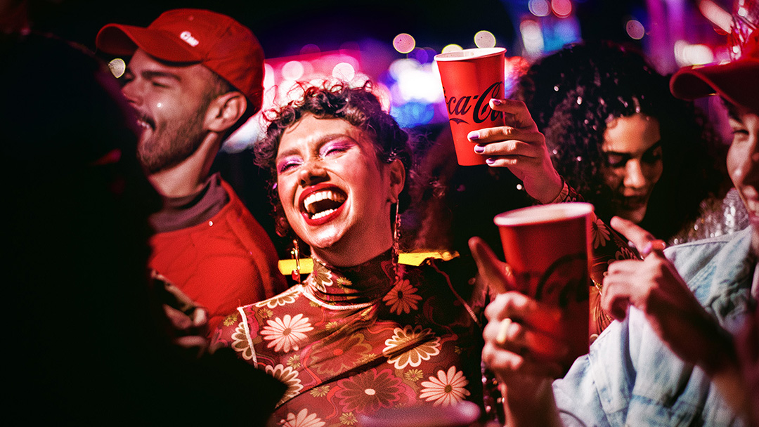 Un grupo de amigos de fiesta por la noche, sosteniendo vasos de Coca-Cola y bailando. Una mujer con el pelo rizado, vistiendo una blusa de patrón floral, está en primer plano riéndose alegremente, rodeada de amigos en un entorno animado y colorido.