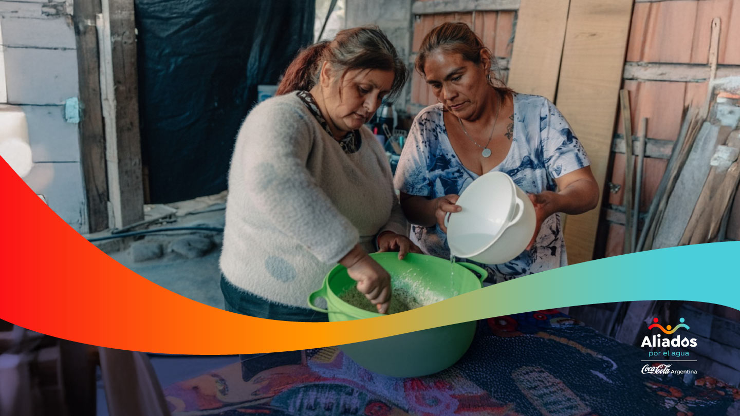 aliados_por_el_agua_ar_desktop.jpg Dos mujeres de mediana edad, preparan alimentos en un entorno doméstico rústico. Una mezcla en un bowl verde mientras la otra vierte agua de un recipiente blanco. En la esquina inferior derecha, el logo 'Aliados por el agua' de Coca-Cola Argentina.