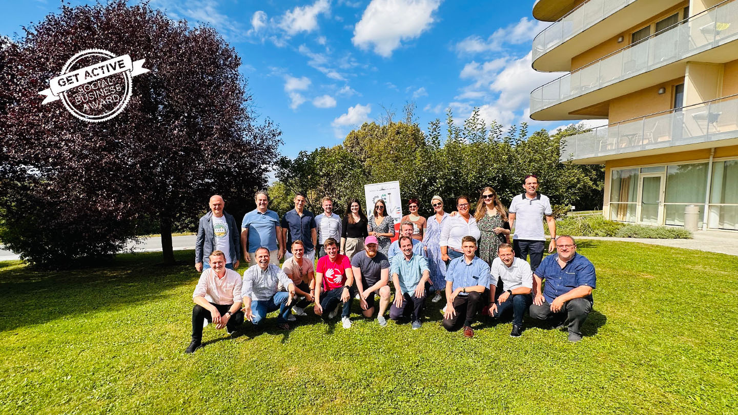 Die 10 Finalist:innen des Get active Social Business Awards mit den GASBA Stakeholders der WU Wien, DerStandard und Coca-Cola Österreich posieren für ein Gruppenfoto auf einer Wiese vor einem Thermenhotel.