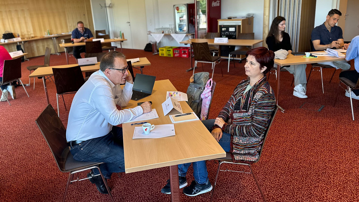 Ein Mann (links) und eine Frau (rechts) sitzen sich an einem schmalen Tisch gegenüber und unterhalten sich.