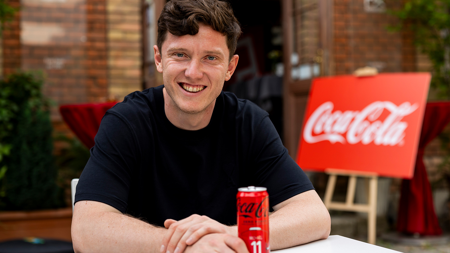 ÖFB-Stürmer Michael Gregoritsch sitzt lächelnd an einem weißen Tisch. Vor ihm steht eine Coca-Cola Zero Sugar Dose mit der Nummer 11, im Hintergrund ist ein rotes Coca-Cola Schild vor einer Backsteinwand zu sehen.