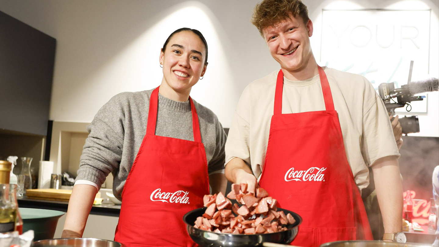 ÖFB-Torfrau Manuela Zinsberger stehet lächelnd in einer Küche. Sie trägt eine rote Coca-Cola-Schürze.