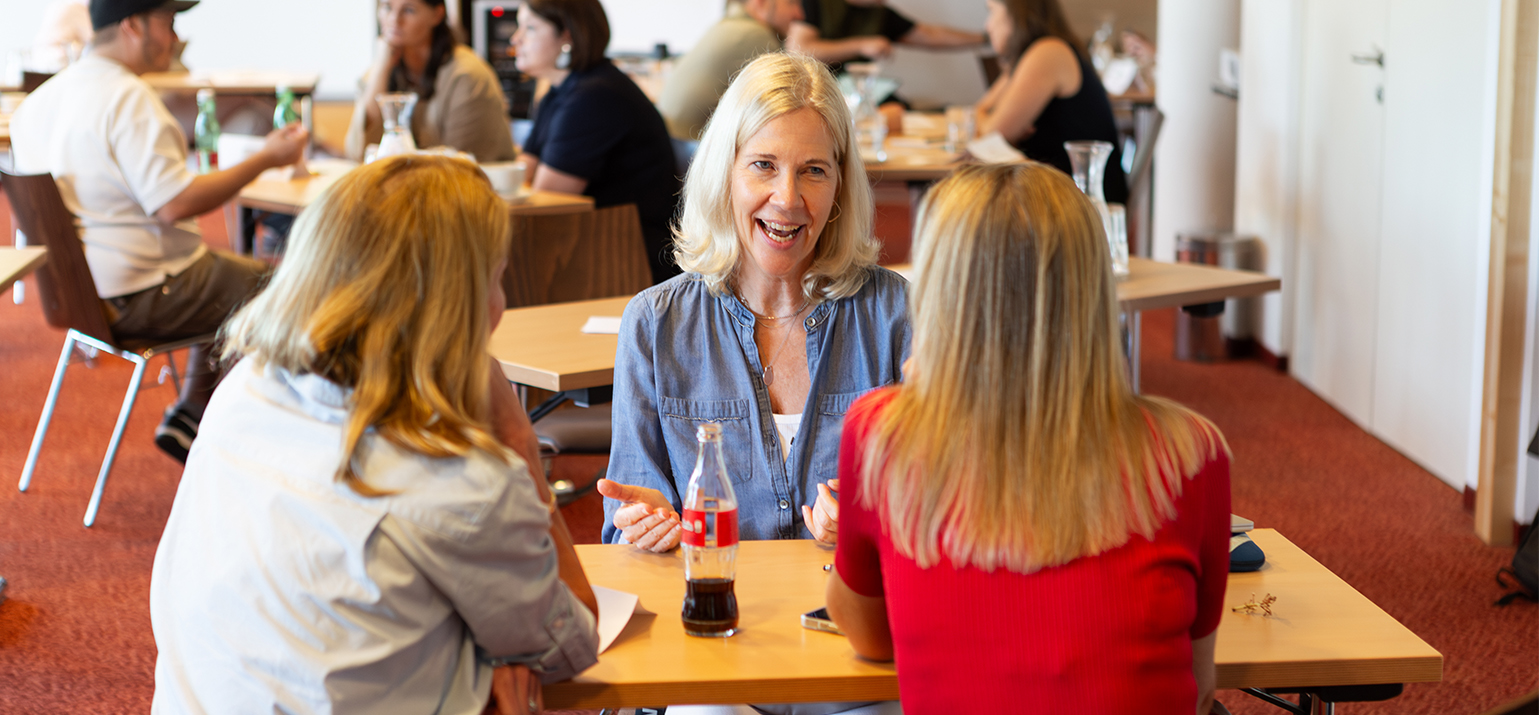 Eine Frau mit blondem Haar im Jeanshemd spricht lächelnd mit zwei anderen Frauen an einem Tisch. Vor ihr steht eine Cola-Flasche. Im Hintergrund sind weitere Teilnehmende an anderen Tischen im Gespräch vertieft.