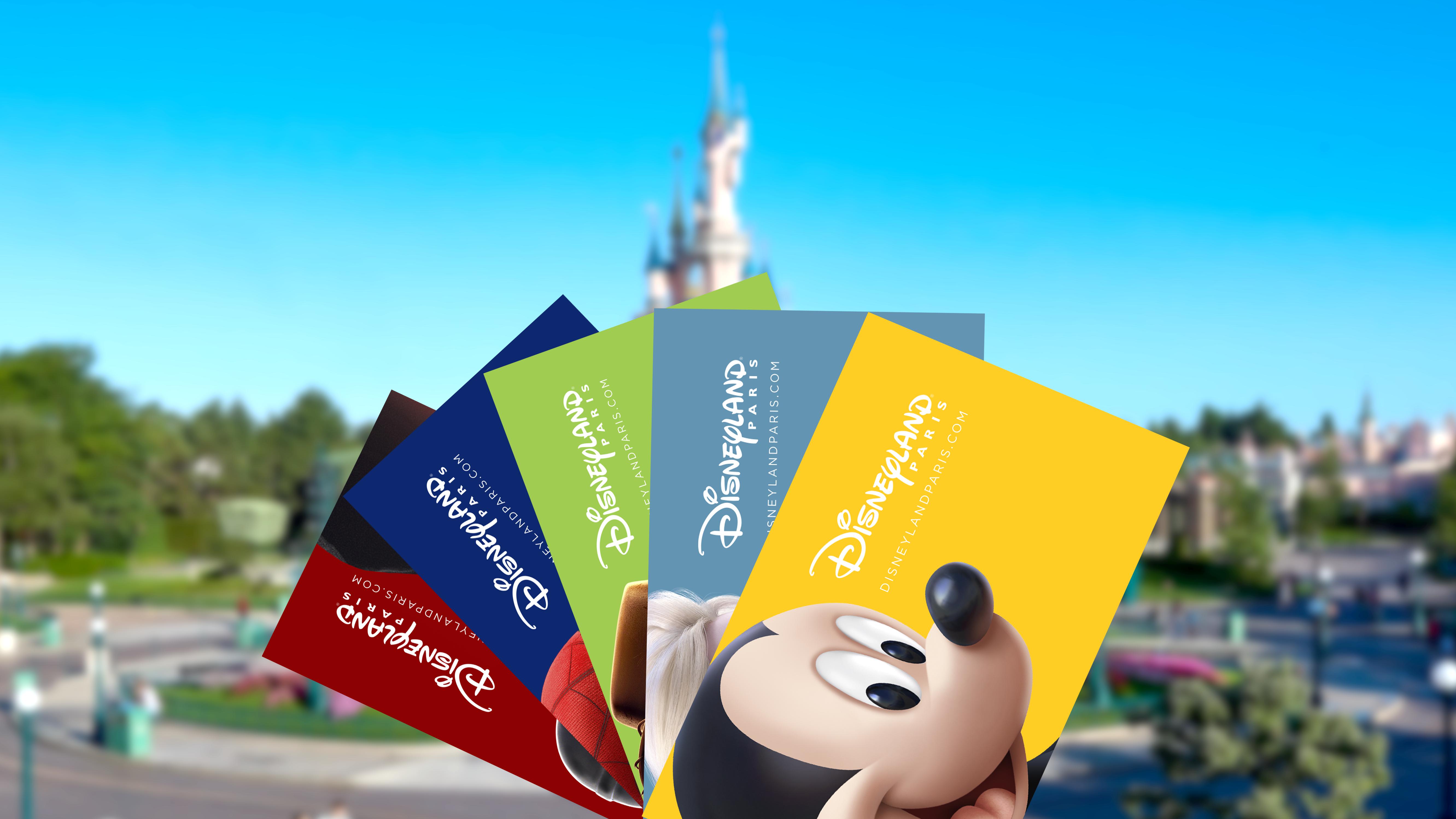 De afbeelding toont vijf Disneyland Paris-toegangspassen in verschillende kleuren en ontwerpen, waaronder een gele kaart met Mickey Mouse en andere kaarten met personages en thema’s van Disney. Op de achtergrond is het iconische kasteel van Disneyland Paris te zien, scherp afstekend tegen een heldere blauwe lucht en omringd door groene beplanting en parkpaden.