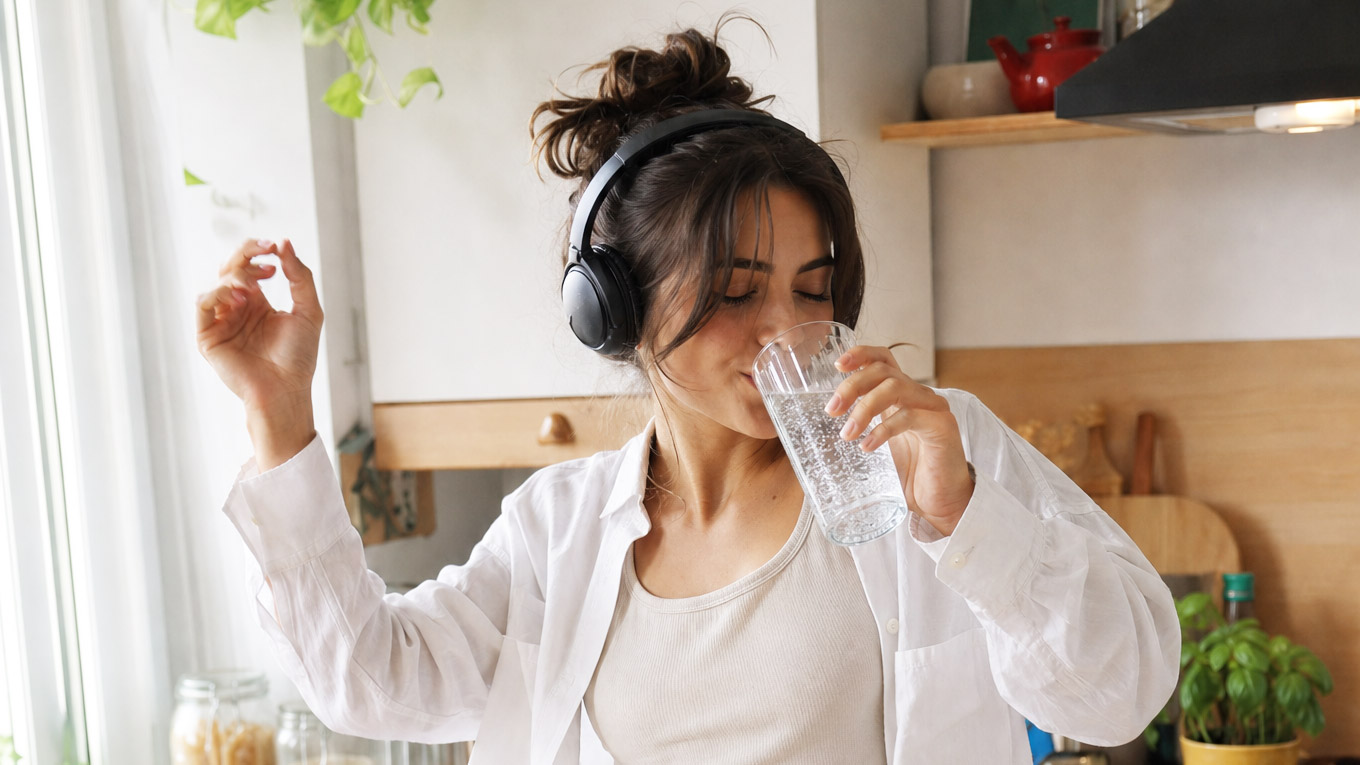 Jonge vrouw in witte outfit en over ear headphones drinkt van een groot glas water in de keuken