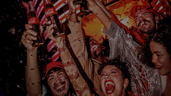 Grupo de amigos celebrando alegremente con botellas de Coca-Cola, rodeados de confeti y luces vibrantes.