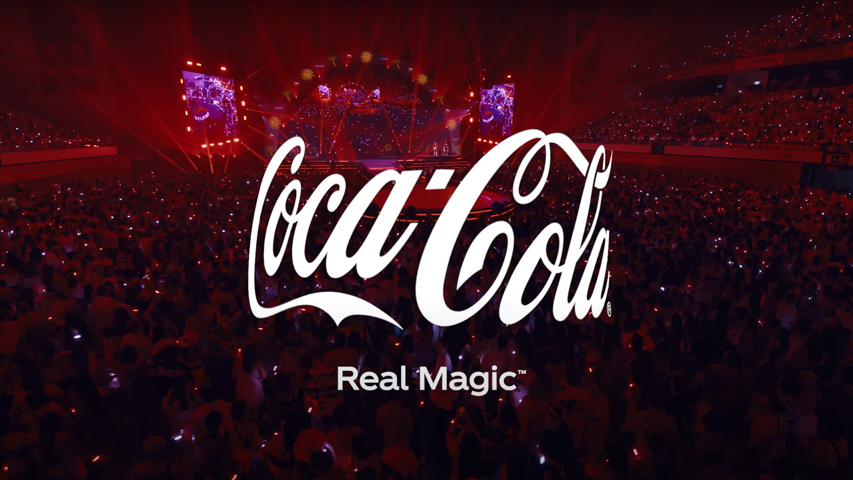 Plano general de un concierto con el logo de Coca-Cola en color blanco superpuesto, con la frase Real Magic debajo del logo.