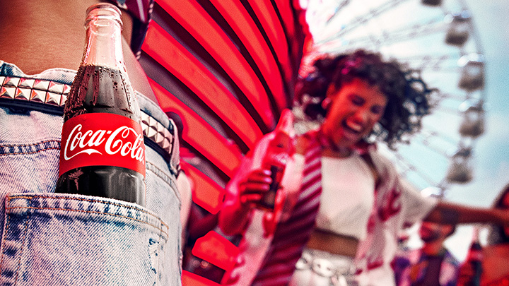 Uma garrafa de Coca-Cola no bolso de uma calça jeans com uma cena de festival alegre ao fundo.