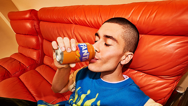 Um jovem está deitado em um sofá de couro laranja vibrante, bebendo uma lata de Fanta. Ele está vestindo uma camiseta azul com um desenho amarelo.