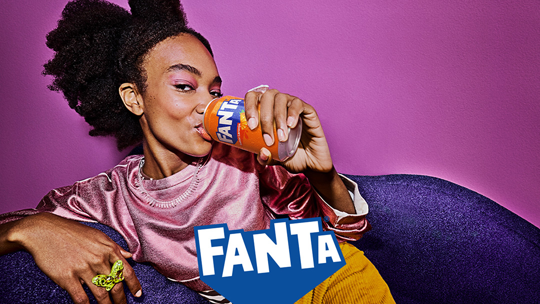 Uma mulher está sentada em um sofá roxo, bebendo uma lata de Fanta. Ela está vestindo uma blusa rosa e calças amarelas. O fundo é uma parede rosa. O logotipo da Fanta está visível na frente do sofá.