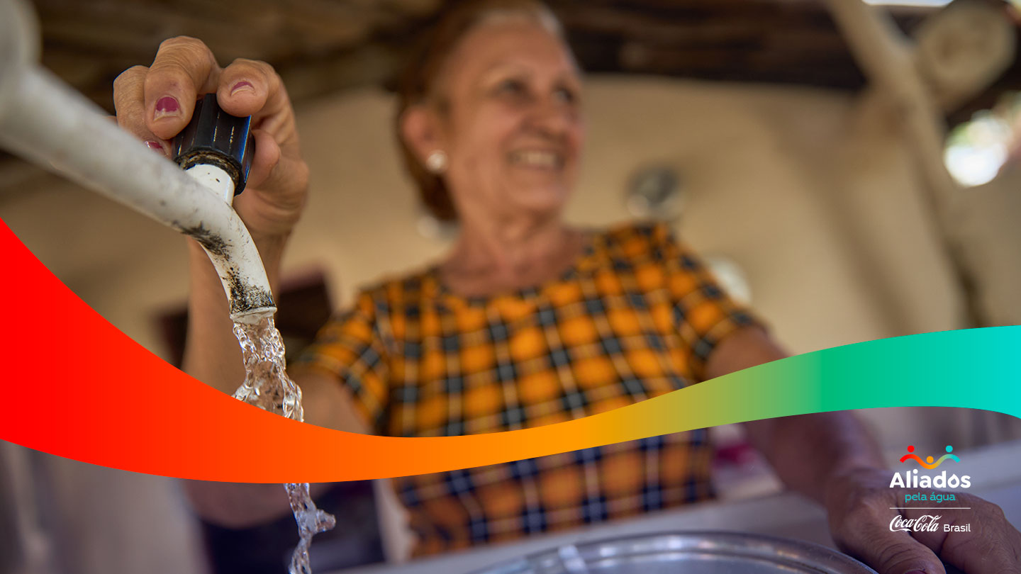 Mulher sorridente abrindo uma torneira de onde flui água limpa, simbolizando o acesso à água potável. No canto inferior direito, vê-se o logo 'Aliados pela Água' junto ao da Coca-Cola Brasil.