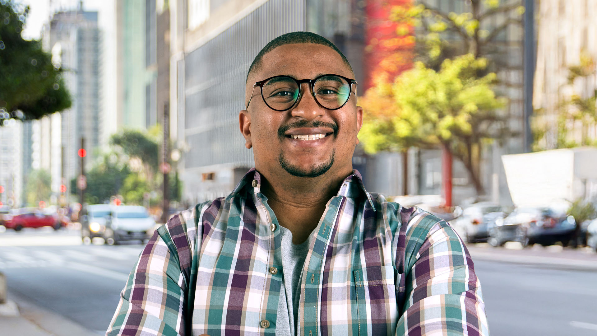 Um homem negro de óculos e camisa xadrez sorrindo em uma rua movimentada da cidade.