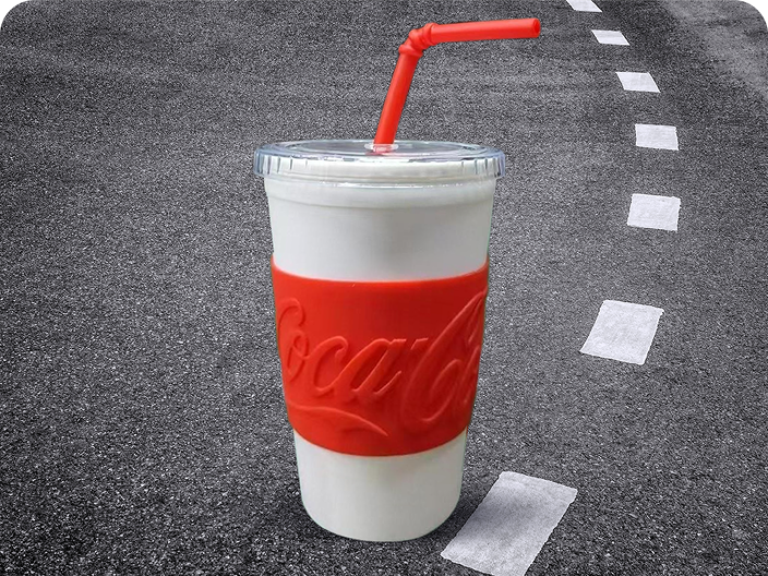 A white tumbler with a red Coca-Cola sleeve and straw, a prize for the Road Trip contest, shown on an asphalt background.