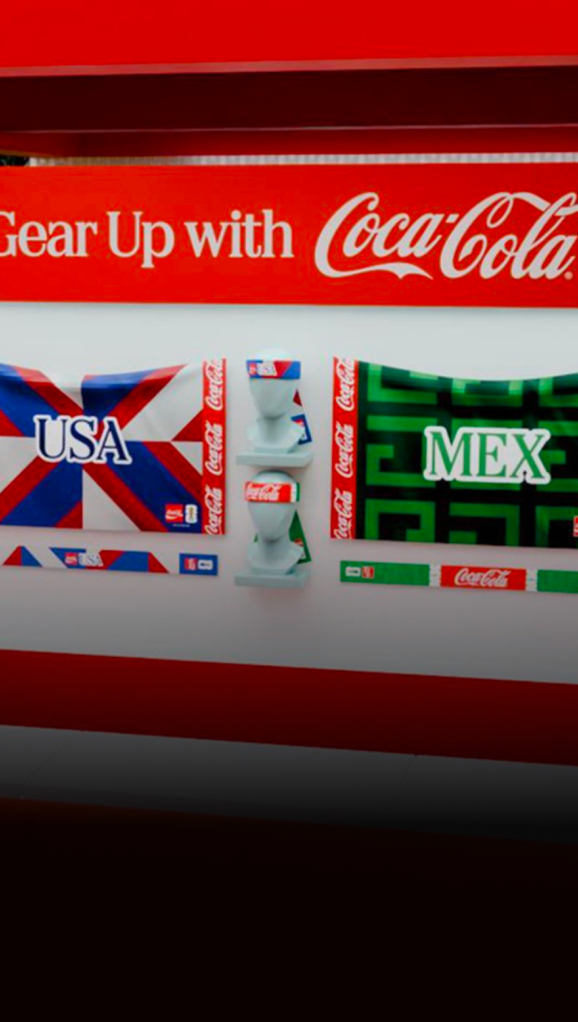 Coca-Cola Fan Zone gear wall with USA and MEX team scarves, headbands and merch displayed under Gear Up with Coca-Cola signage.