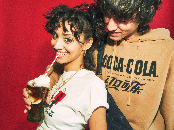 A young woman drinking a cola float through a striped straw stands in front of a young man wearing a tan hoodie printed with the "Coca-Cola" logo, against a red background.