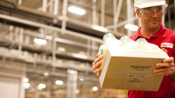 A factory worker with a red uniform and safety equipment carrying a carton box with products