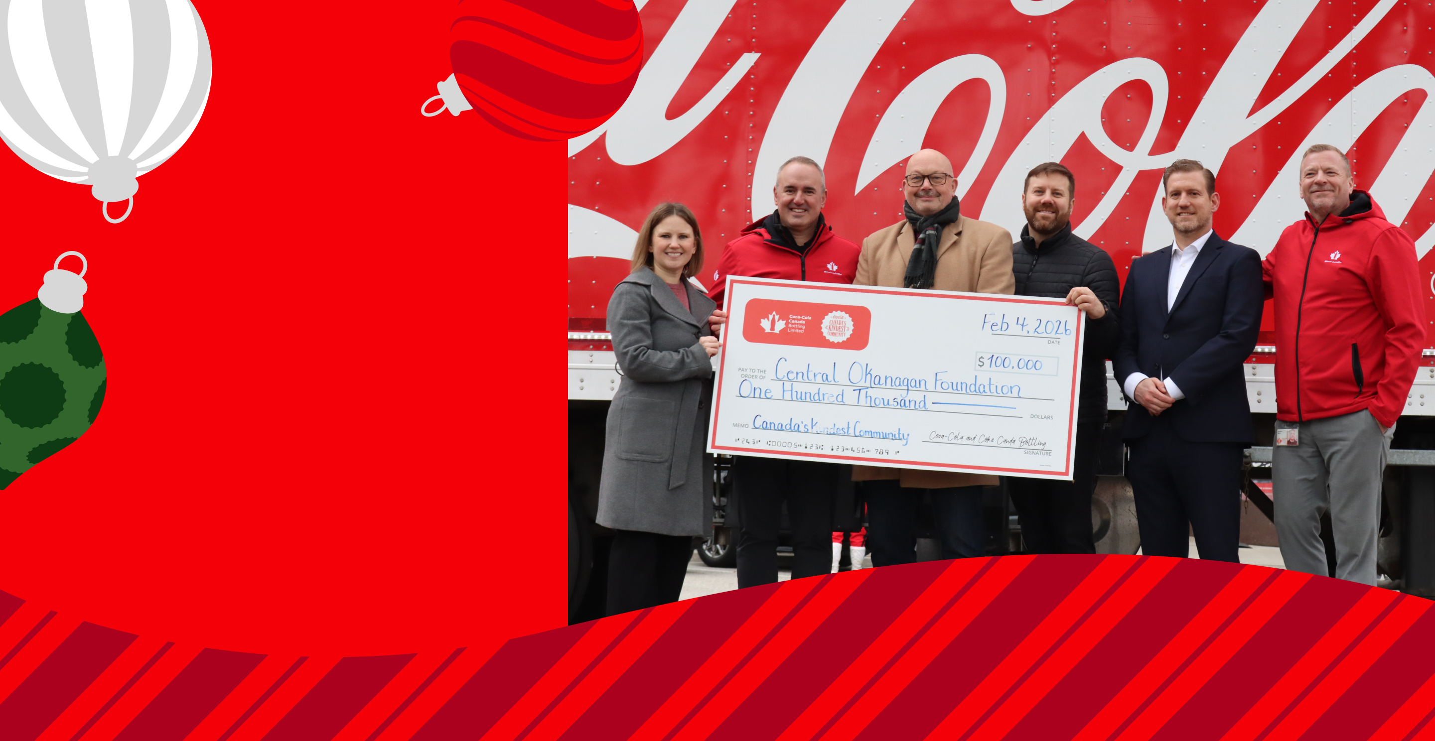 Personnes se tiennent devant un camion de livraison Coca-Cola en tenant un chèque géant de 100 000 $ daté du 4 février 2026, payable à la Fondation Central Okanagan, avec la mention « Canada's Kindest Community », entourées de motifs d'ornements de Noël.