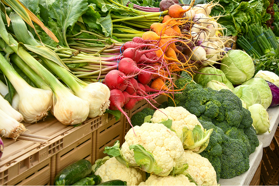 Un assortiment de légumes frais — dont du fenouil, des radis, du chou-fleur, du brocoli et des betteraves — disposés dans des cageots à un étal de marché extérieur.