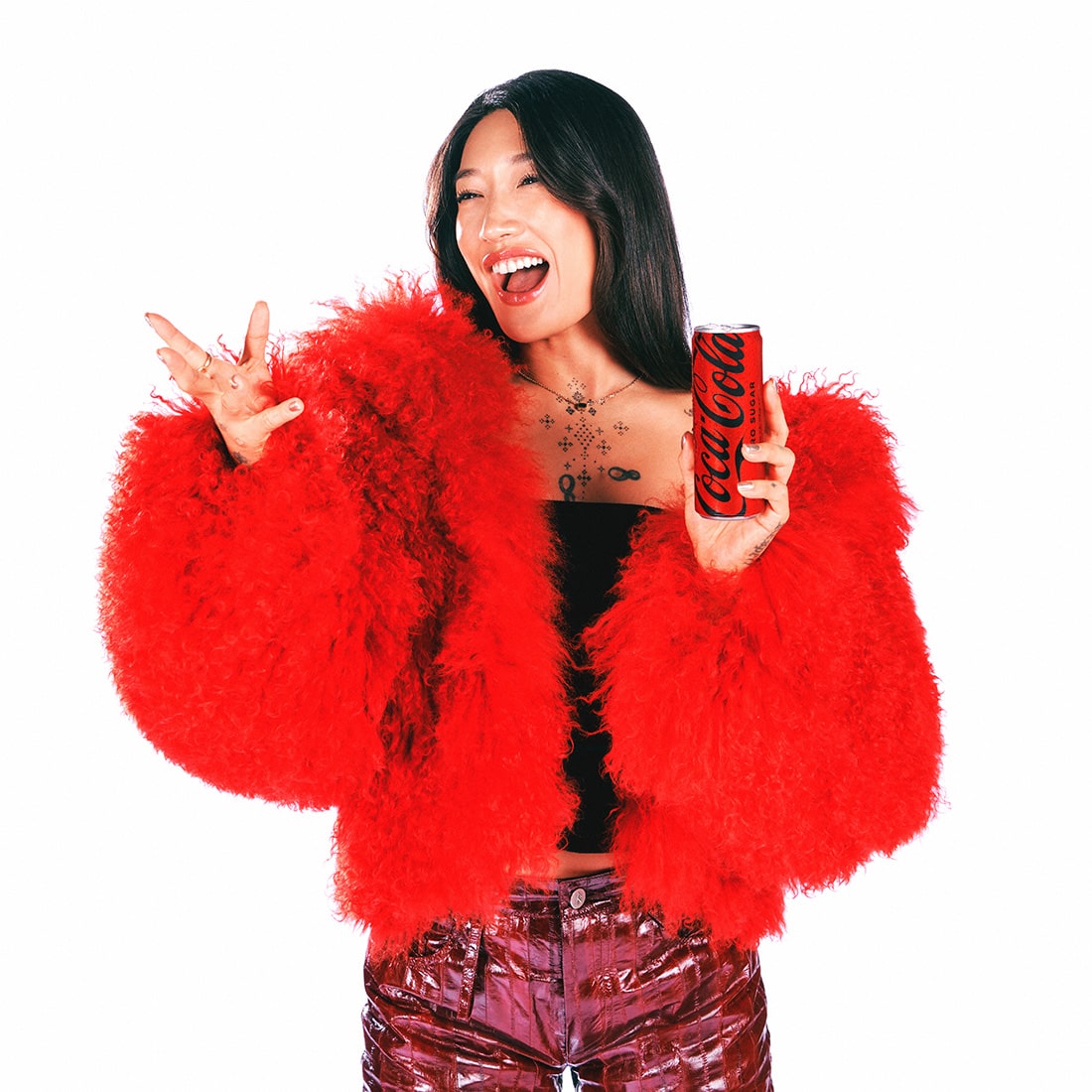 Peggy Gou, with a big smile and wearing a red furry coat, holds a limited edition Coca-Cola can while making a joyful hand gesture.