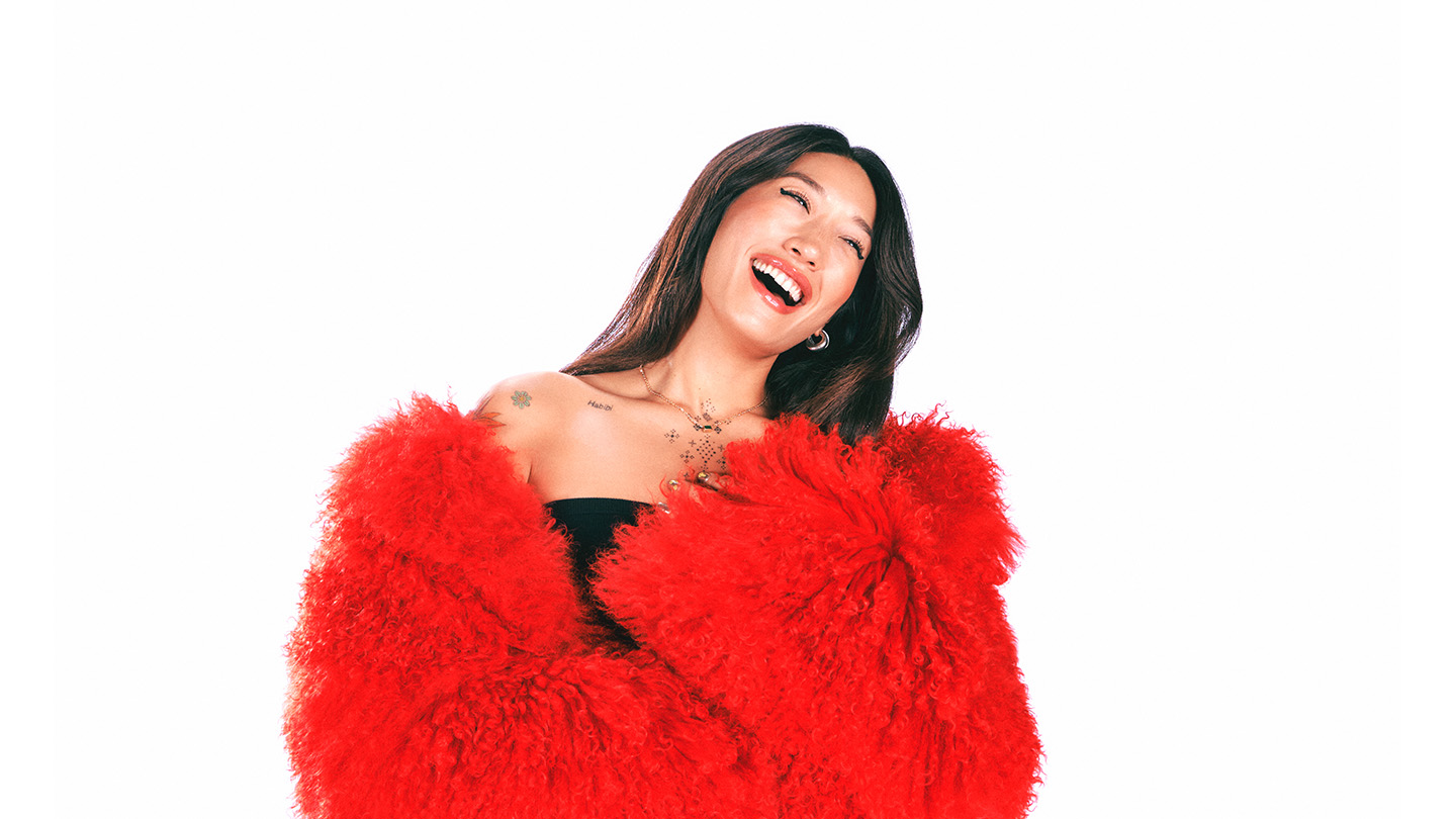 Peggy Gou laughing, wearing a red fur jacket, against a white background.