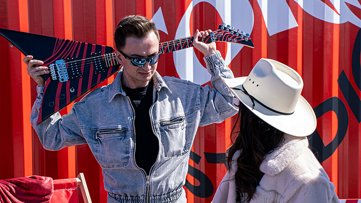 A man holding a guitar behind his head, talking to a woman wearing a cowboy hat, with a vibrant Coke Studio backdrop.