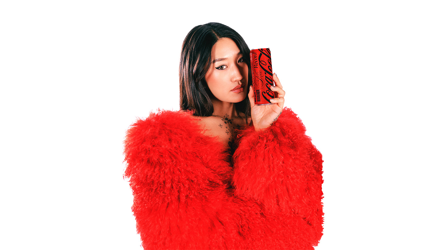 Peggy Gou, a young woman with dark, long hair, wearing a bright red furry coat, holds a limited edition Coca-Cola can close to her face, looking at the camera with a serene expression.