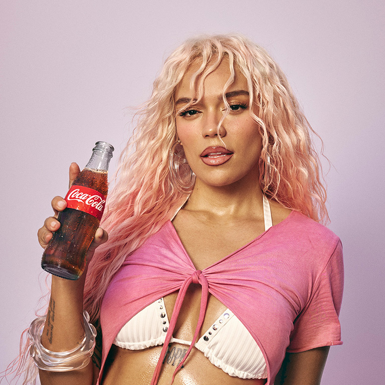 Karol G posing with a Coca-Cola bottle against a neutral background.