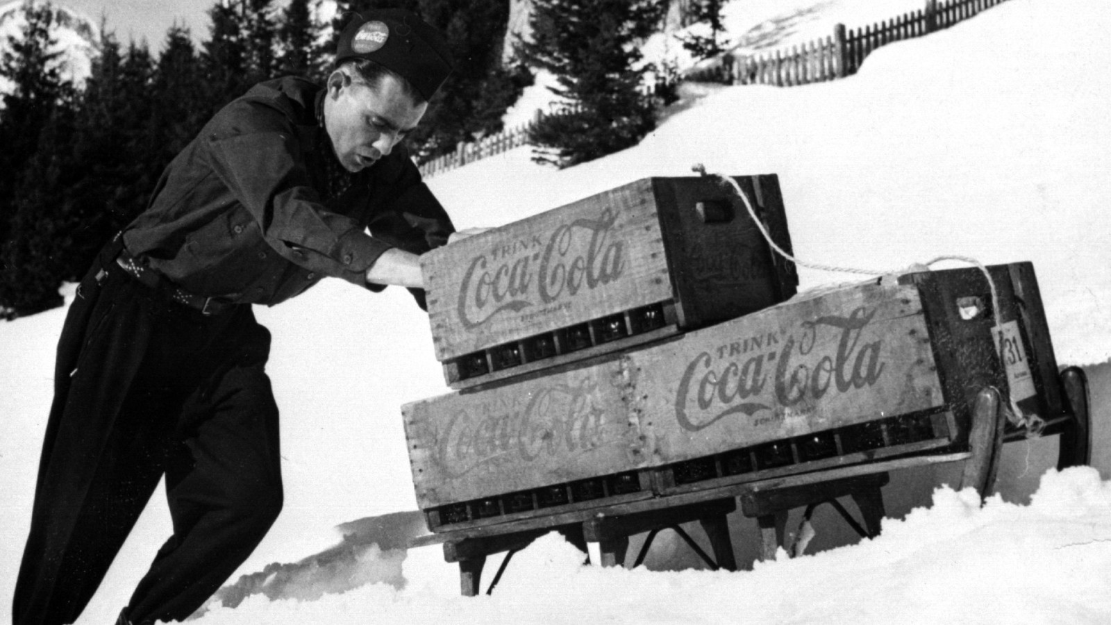 Historische Aufnahme eines Mannes im Schnee mit einem Schlitten, der mit Kisten von Coca-Cola beladen ist.