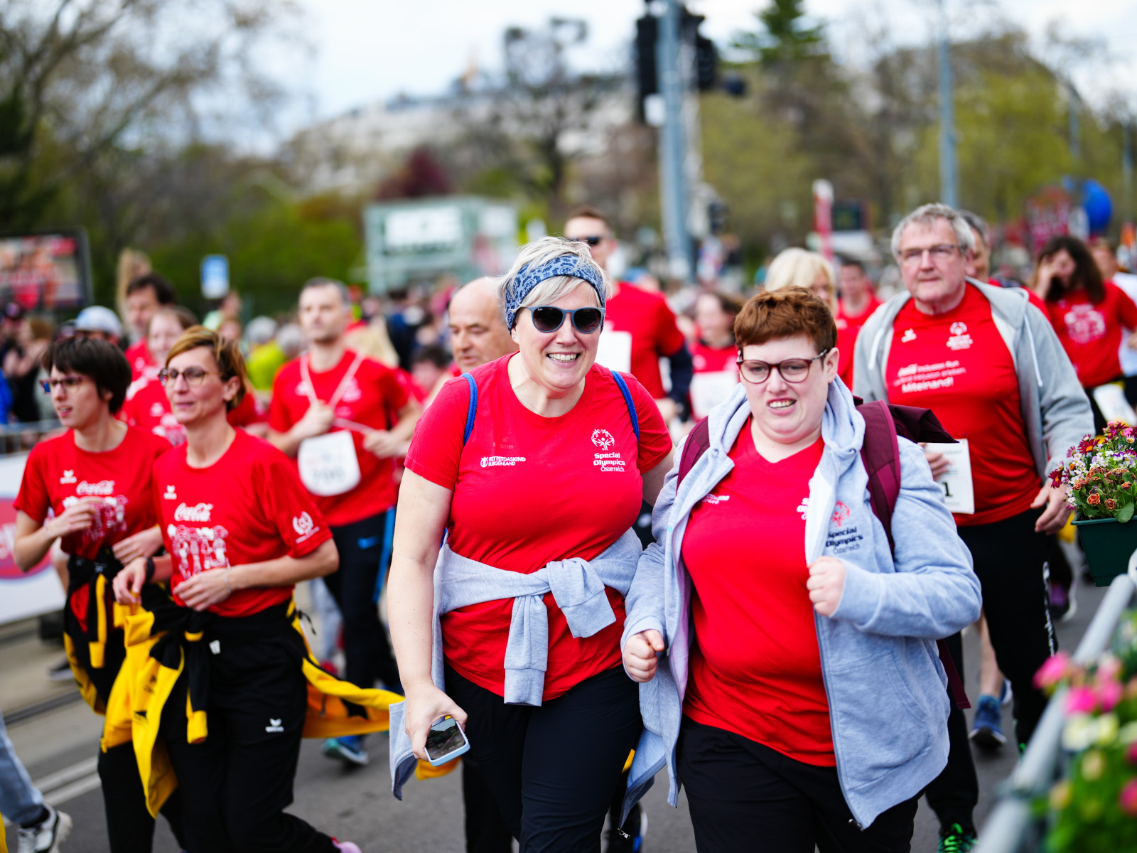 Zwei Läuferinnen am Coca-Cola Inclusion Run 2025 in Wien schauen in die Kamera, während hinter ihnen weitere Läuferinnen die Strecke bestreiten.