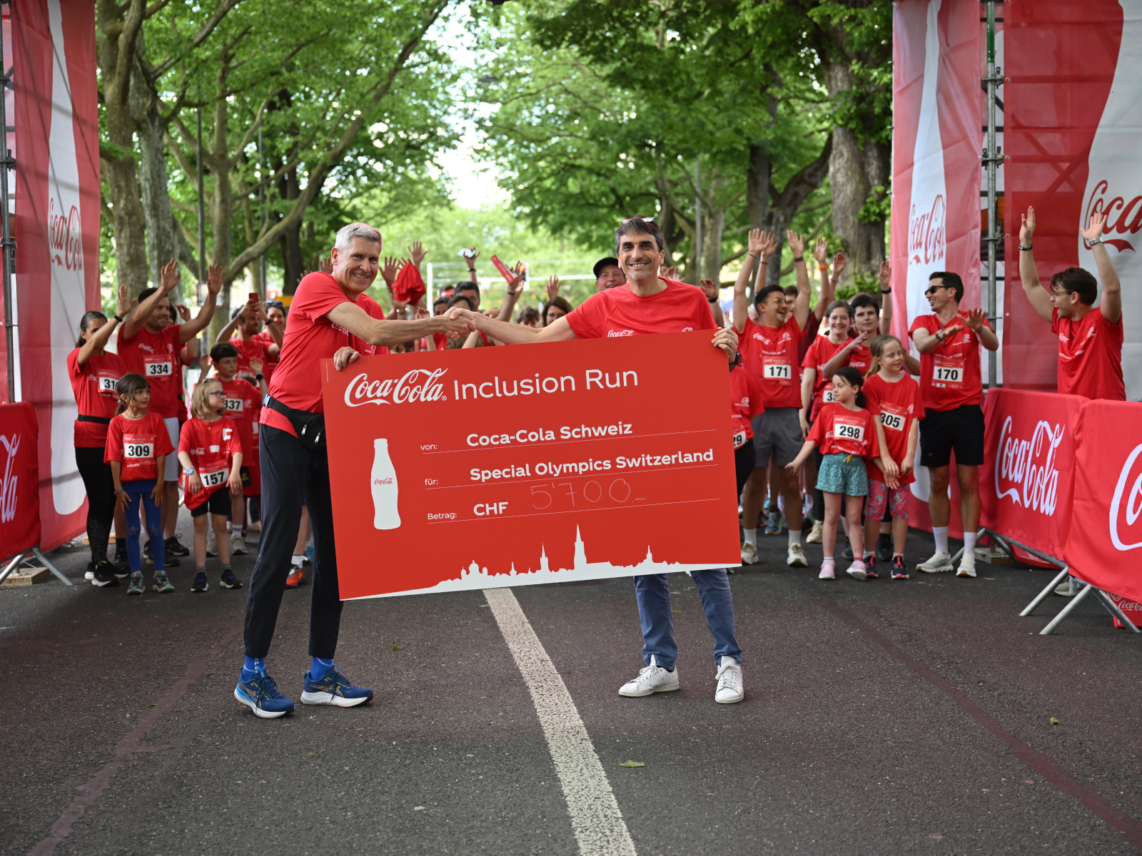 Zwei Männer halten beim Start der Laufstrecke ein Plakat mit einer Spende hoch und schütteln sich die Hand, im Hintergrund machen die Läuferinnen und Läufer eine Laola-Welle.