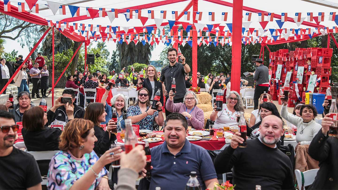 Una multitud de personas sonríe y brinda con botellas de Coca-Cola en un evento al aire libre. La escena está decorada con guirnaldas rojas, blancas y azules, y hay mesas con comida.