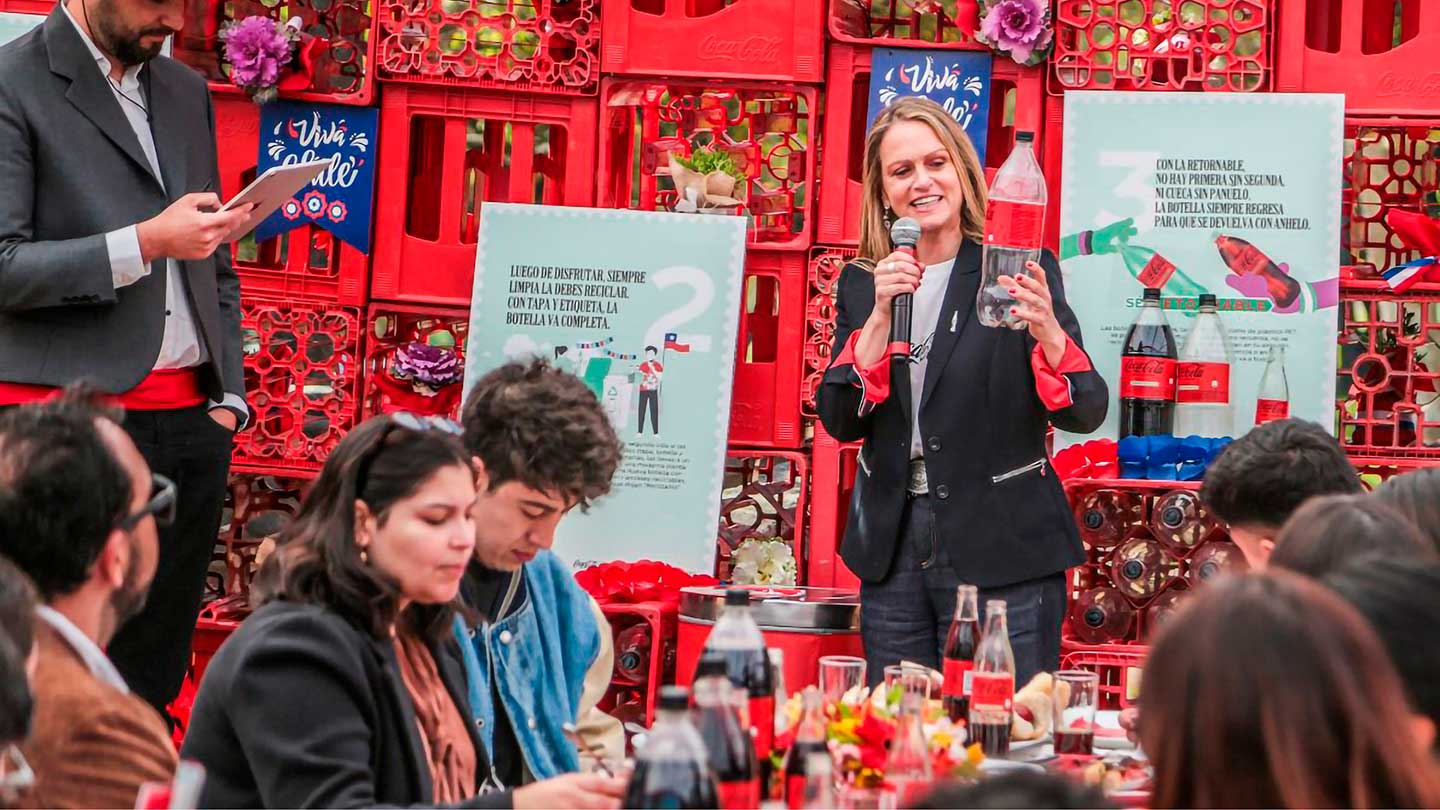 Una mujer sonriente con un micrófono en la mano sostiene una botella de Coca-Cola, dirigiéndose a un grupo de personas sentadas alrededor de una mesa. Detrás de ella, hay una pared decorada con cajas rojas de Coca-Cola y carteles informativos.