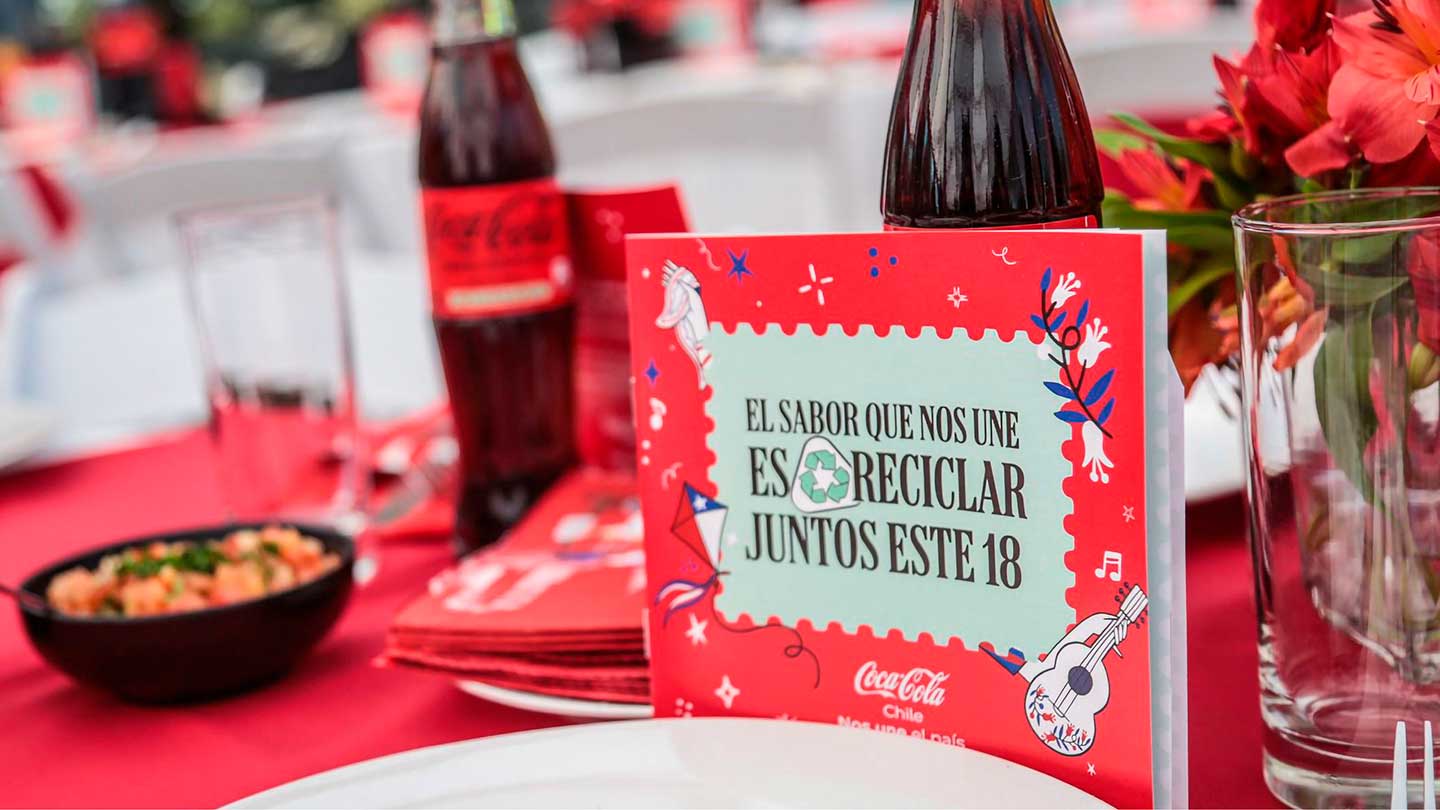 Una mesa decorada con un mantel rojo, botellas de Coca-Cola, un plato con comida y un cartel que dice "EL SABOR QUE NOS UNE ES RECICLAR JUNTOS ESTE 18".