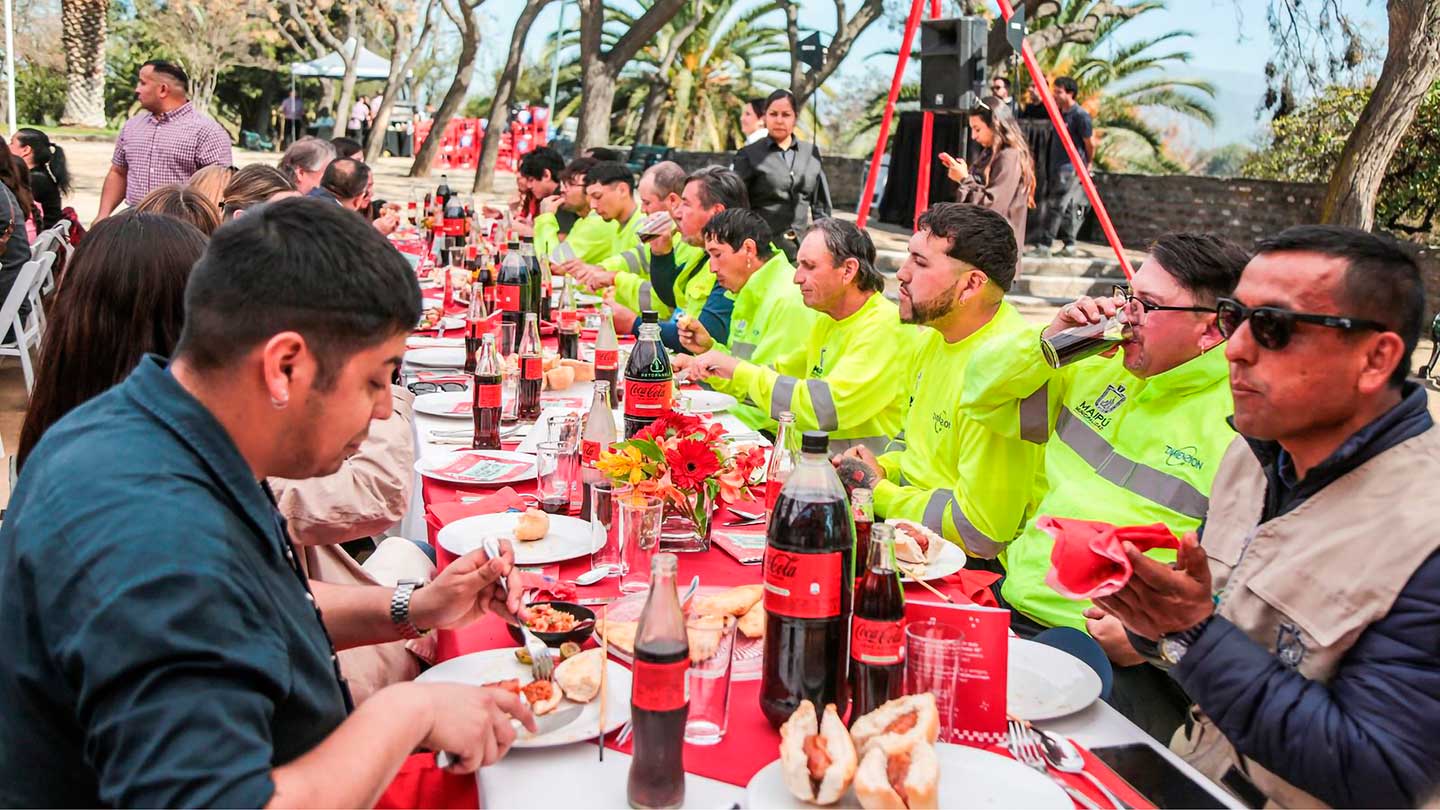 Un grupo de personas, algunas con chalecos de seguridad, comen y beben Coca-Cola en una larga mesa al aire libre.