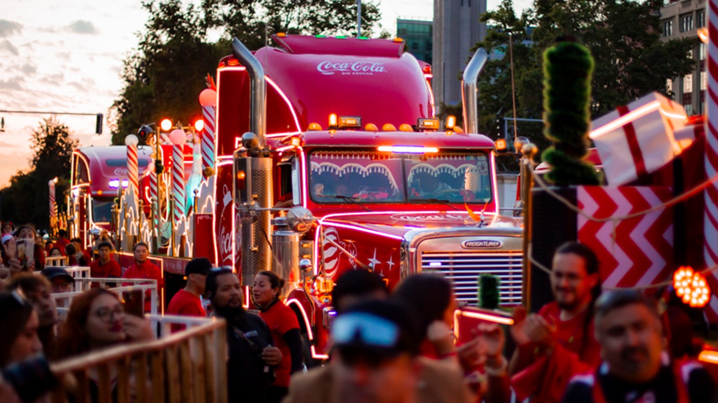 Un camión decorado con luces navideñas brillantes y el logo de Coca-Cola sin azúcar, rodeado de personas celebrando en las calles durante la Caravana Navideña.