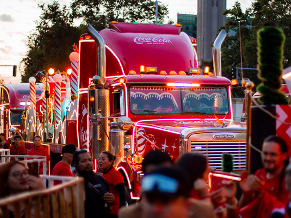 Un camión decorado con luces navideñas brillantes y el logo de Coca-Cola sin azúcar, rodeado de personas celebrando en las calles durante la Caravana Navideña.