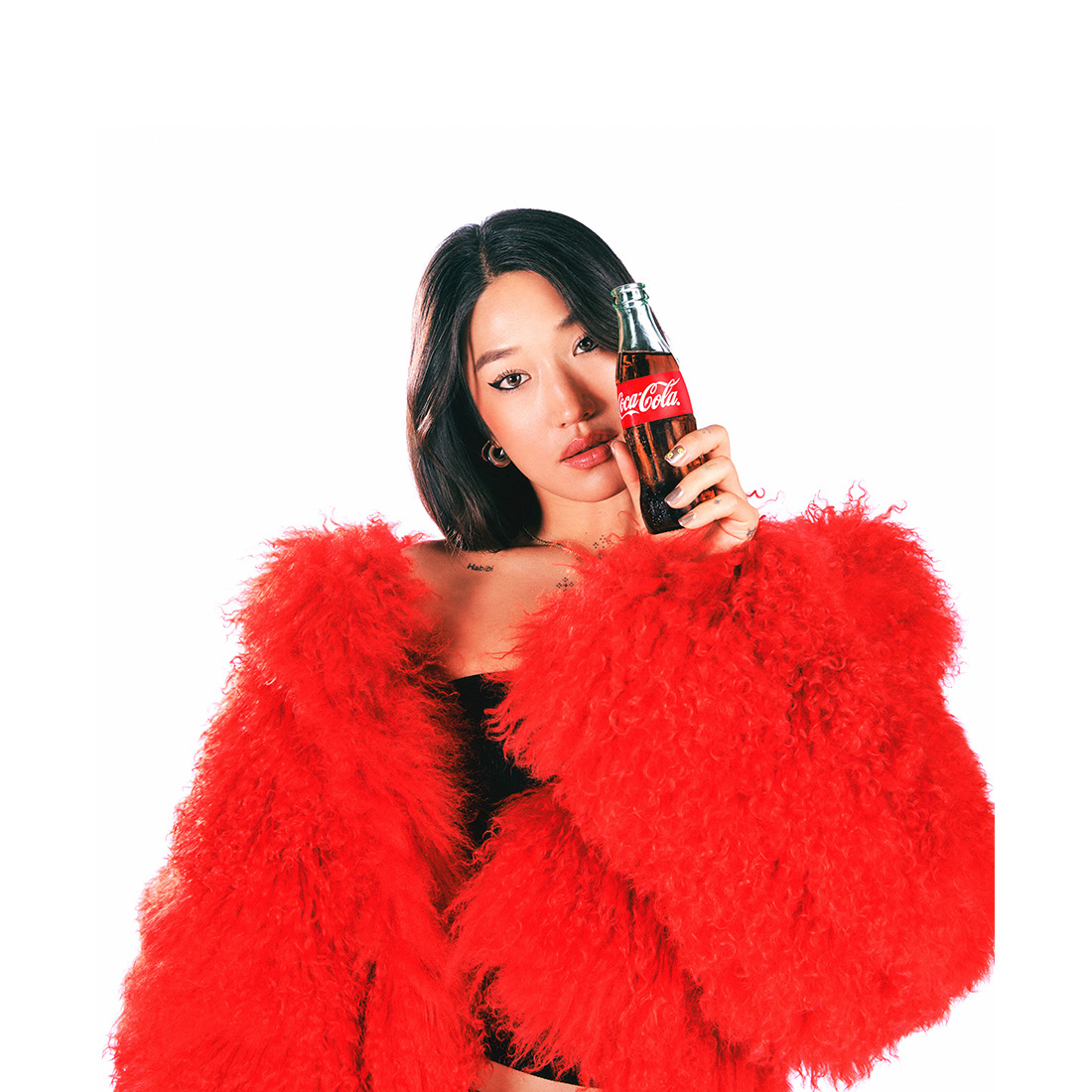 Peggy Gou, una mujer joven con cabello oscuro y largo, viste un abrigo de peluche rojo brillante y sostiene una botella de Coca-Cola de vidrio cerca de su rostro, mirando a la cámara con una expresión tranquila.