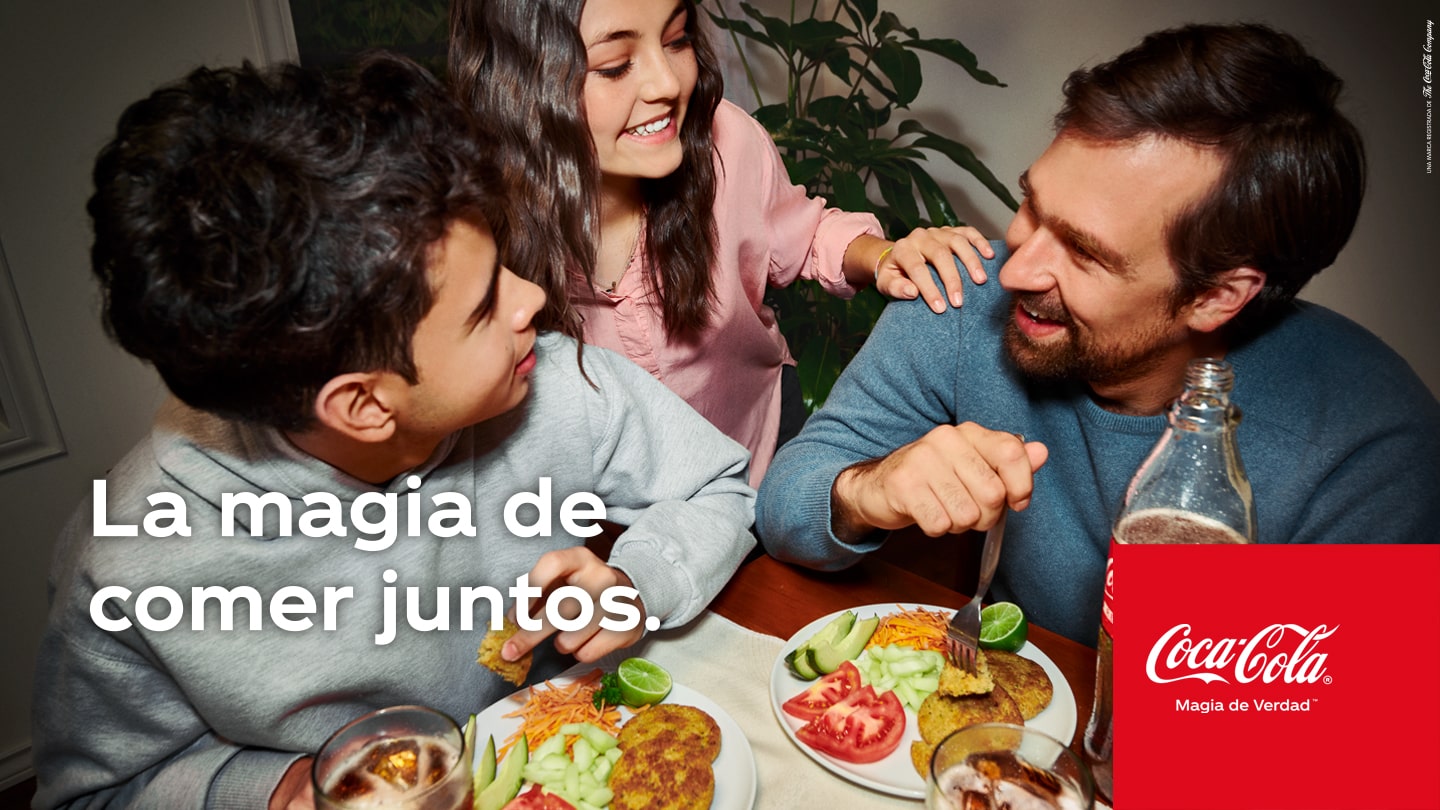 Una familia disfrutando de una comida en la mesa riendo y compartiendo una Coca-Cola. Con un texto sobre el lado izquierdo inferior de la imagen que dice “La magia de comer juntos.” y el logo de Coca-Cola sobre el lado derecho.