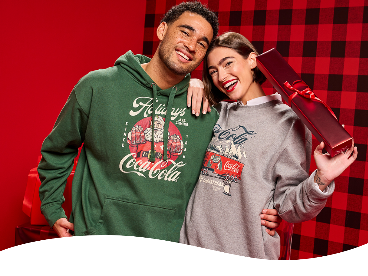 Paar hält ein verpacktes Geschenk in Coca‑Cola‑Weihnachts-Sweatshirts mit Santa‑Motiven.  Santa trinkt Coca‑Cola aus einer Glasflasche vor einem roten Punktmuster-Hintergrund.