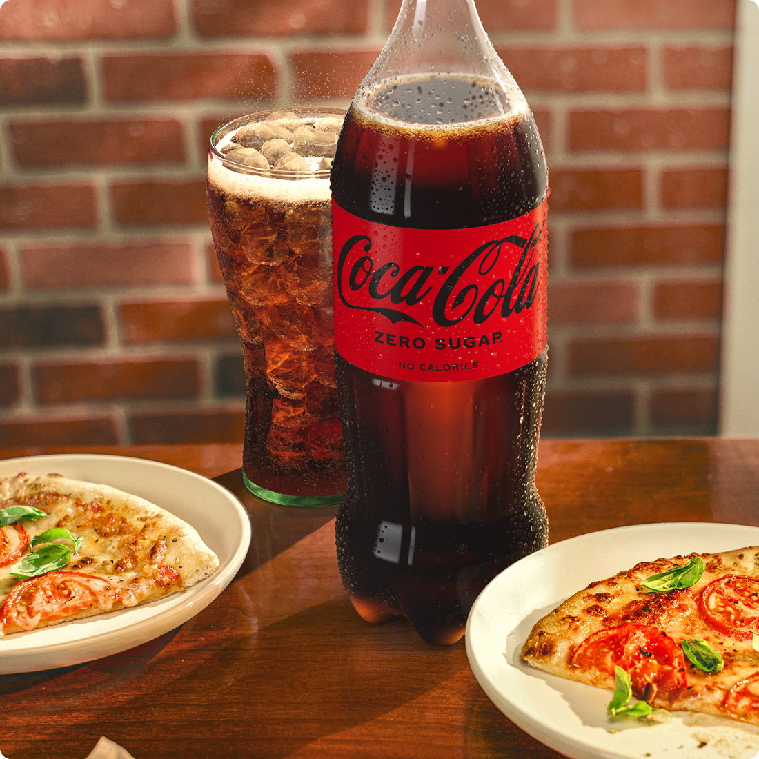 A bottle of Coca-Cola zero sugar and a full glass is on a table. Beside it are two plates of pizza.