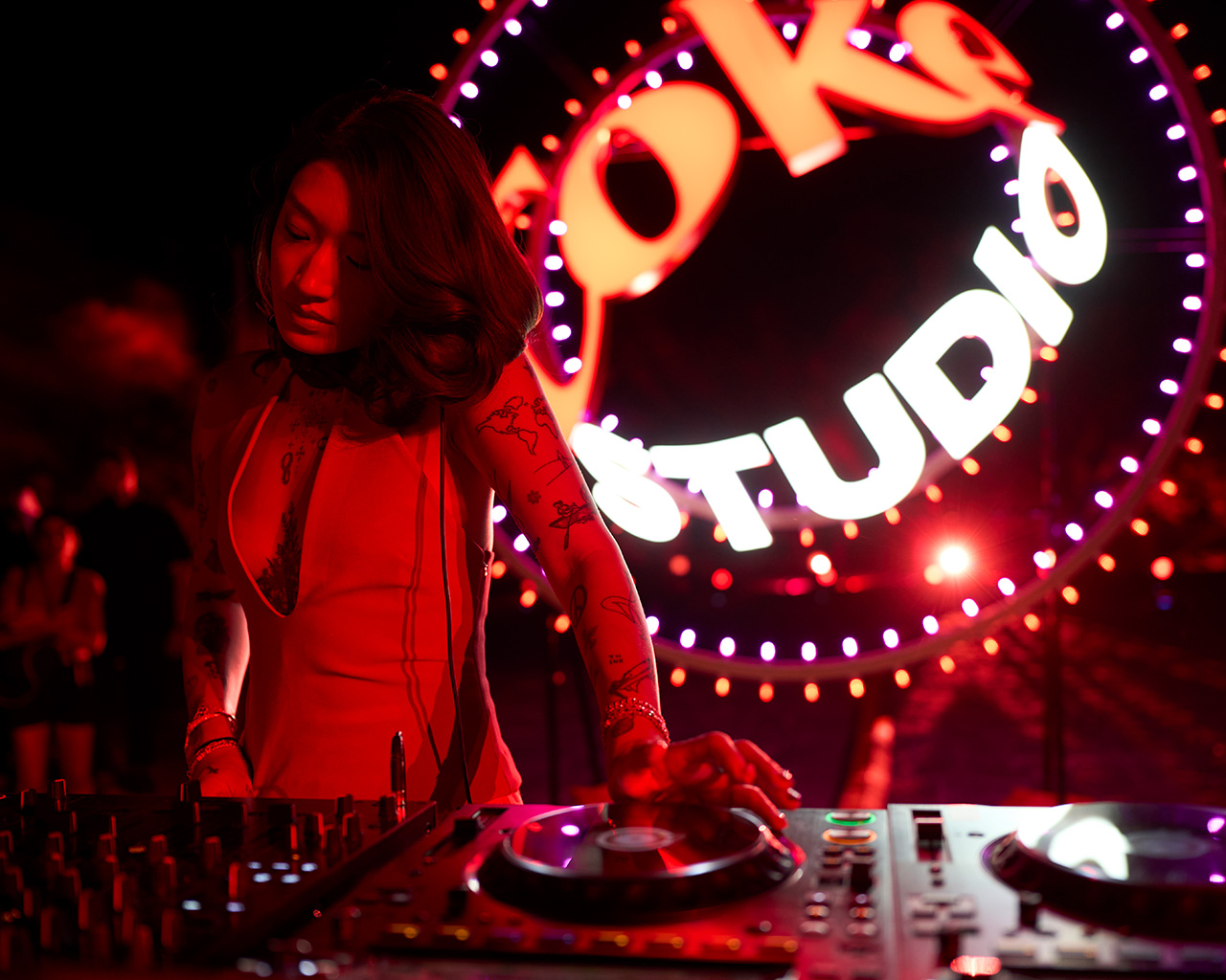 Peggy Gou tocando en vivo en un evento de Coke Studio, con iluminación vibrante en rojo y el logo al fondo. Lleva un vestido rojo y tiene tatuajes visibles en los brazos. Al fondo, hay personas y un ambiente de fiesta y música