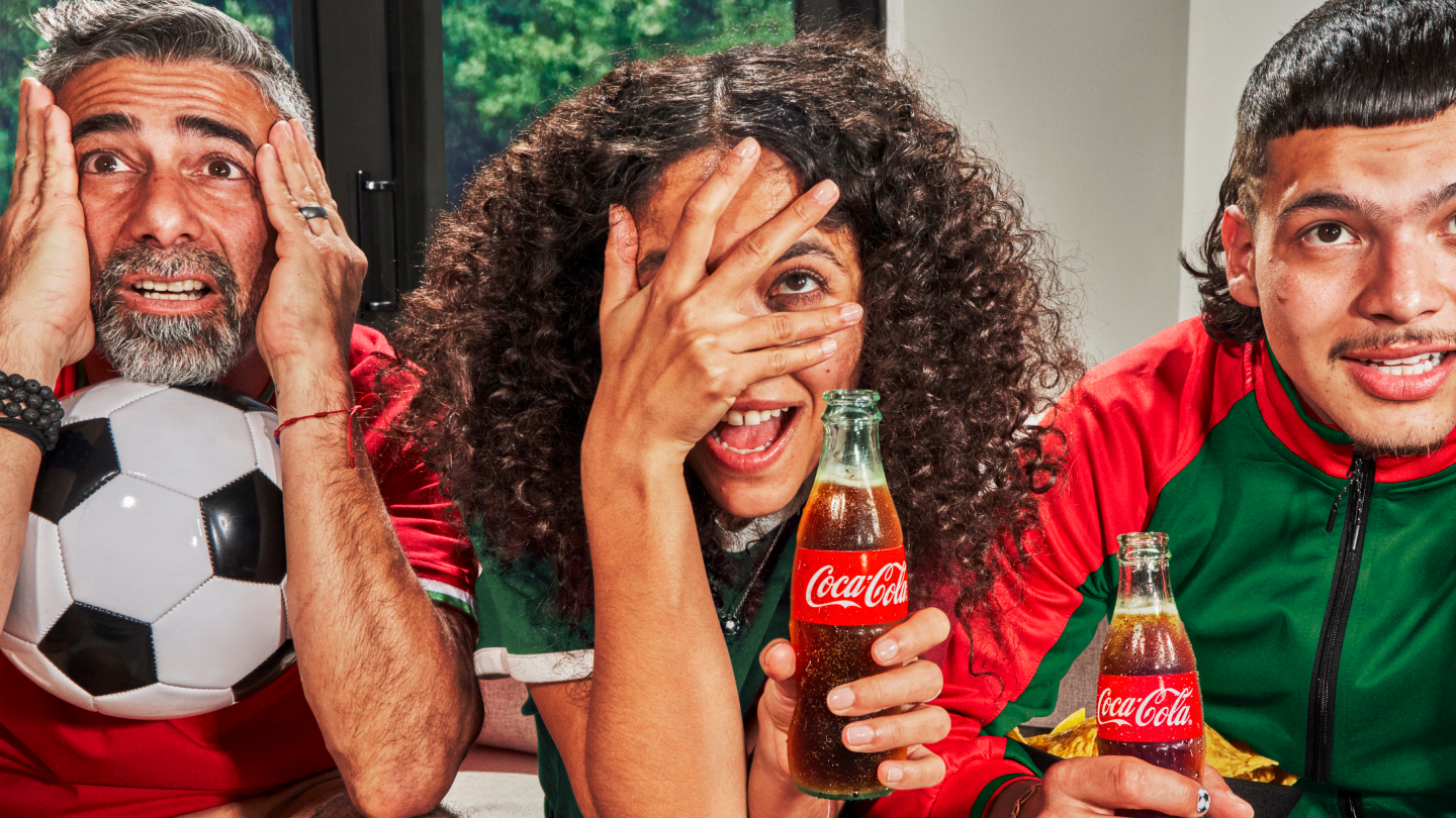 Trois supporters de football portant des maillots rouge et vert applaudissent avec les poings levés ; une écharpe est tenue en l’air et un ballon de football est visible en arrière-plan.