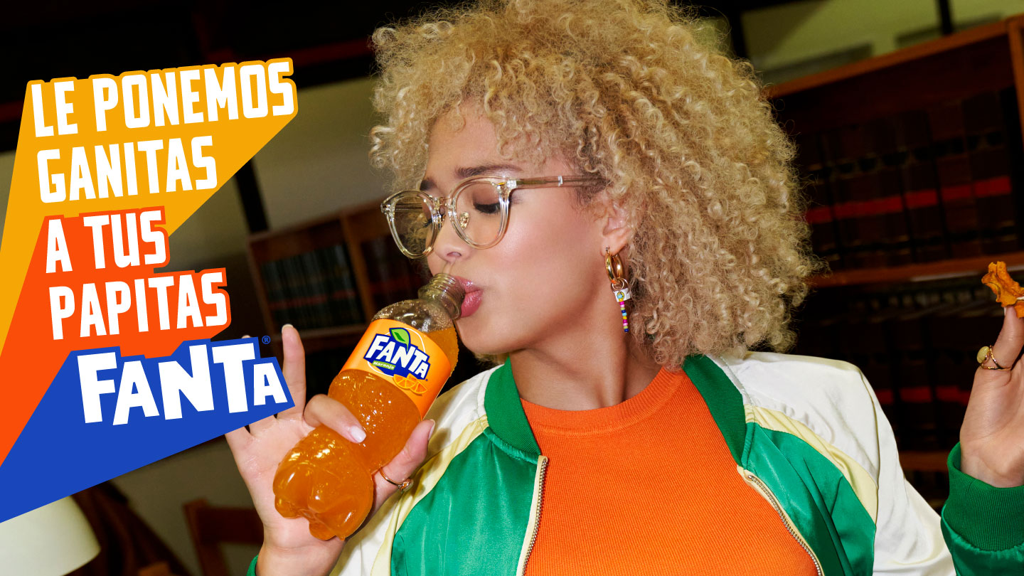 Mujer de pelo enrulado rubio, con anteojos y ropa colorida bebiendo una botella de Fanta sabor naranja con un snack en la otra mano dentro de una biblioteca. A su lado hay un texto que dice “Le ponemos ganitas a tus papitas” y por debajo está el logo de Fanta.