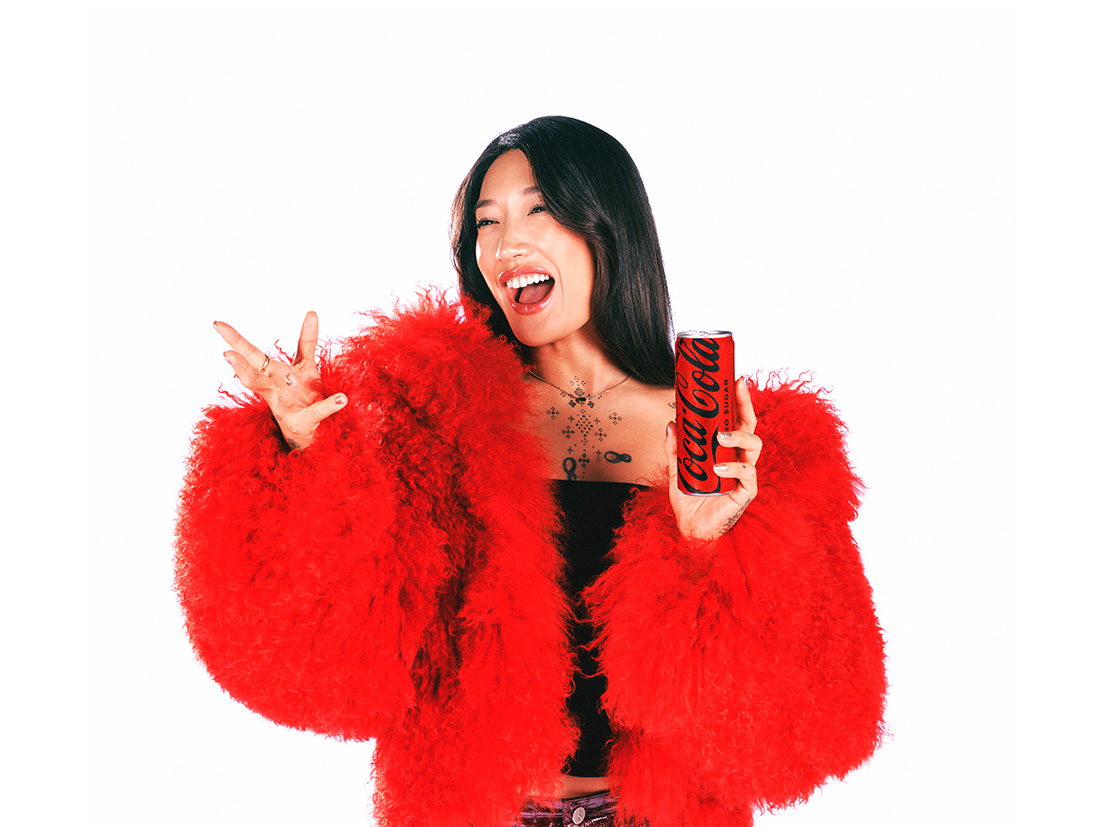 Peggy Gou sosteniendo una lata de Coca-Cola mientras lleva una chaqueta de piel roja, sobre un fondo blanco.