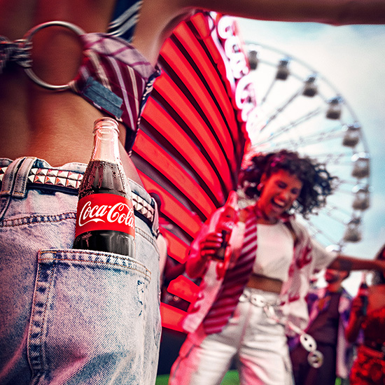 Una botella de Coca-Cola en el bolsillo de un pantalón de mezclilla, con una escena alegre de festival en el fondo.