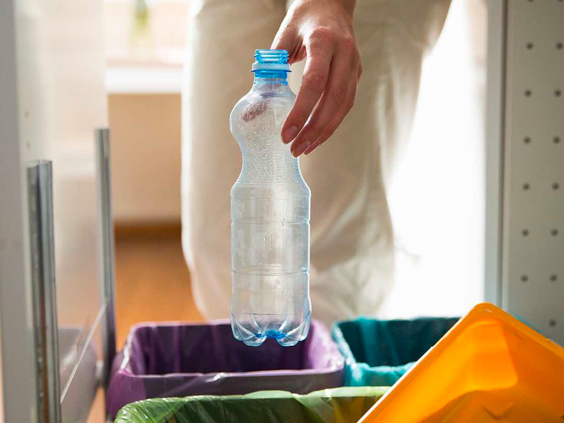 A person putting an empty plastic bottle inside a trash can with a purple plastic bag