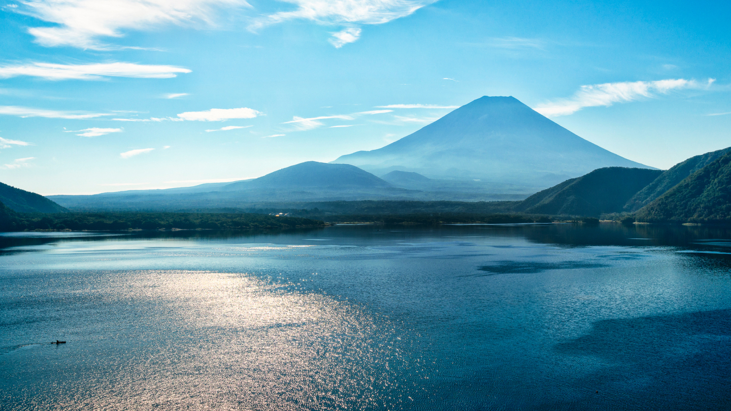 写真：静かな湖が広がり、遠くの山々が見え、晴れ渡った青空に白い雲がいくつか浮かんでいる風景。