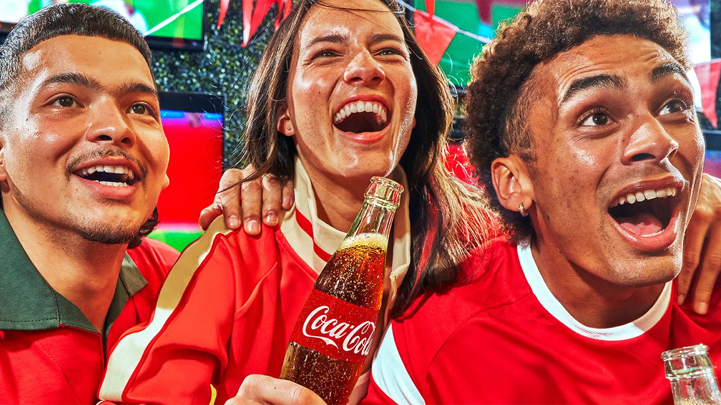 빨간색 유니폼을 입은 축구 팬들이 코카-콜라를 마시며 신난 표정으로 FIFA 월드컵 경기를 응원하는 모습.