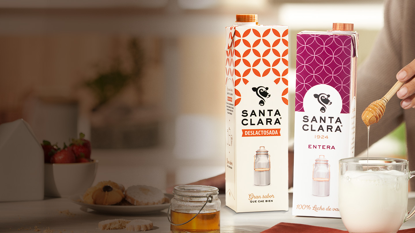 Dos envases de leche Santa Clara, uno con diseño naranja y otro morado, se muestran en una superficie. A la derecha, una mano vierte miel con un cucharón en un vaso de leche. También se ve un frasco de miel y elementos borrosos de fondo.